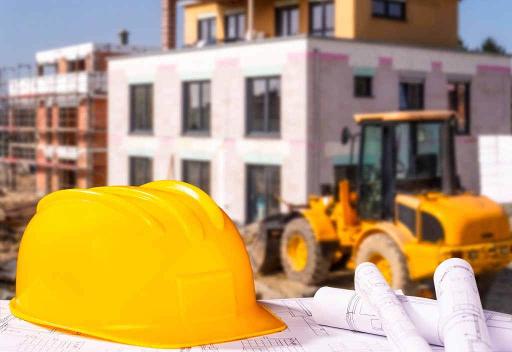 casque jaune avec plans de bâtiments en papier en premier plan, tracteur avec bâtiment en second plan